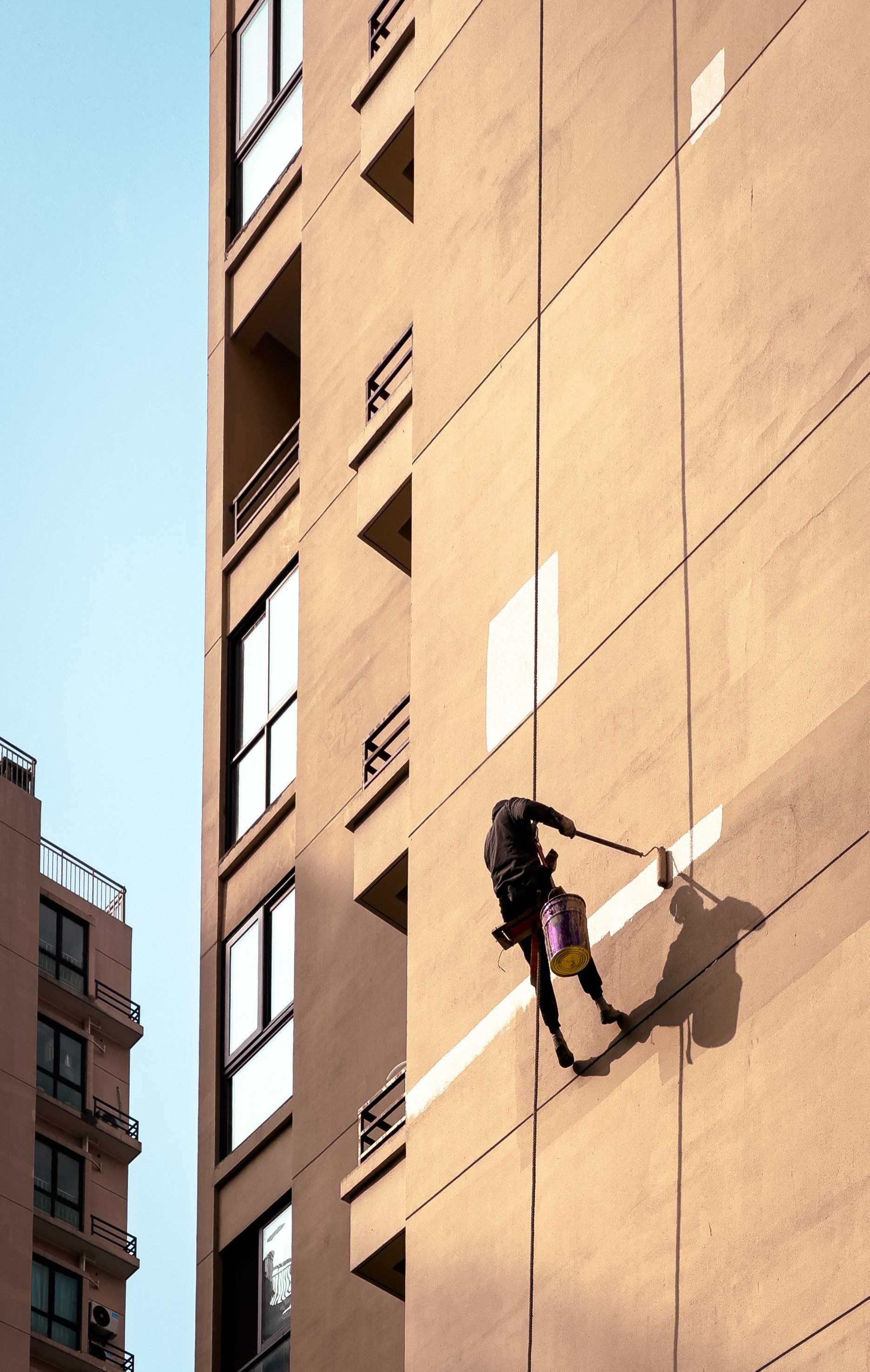 Man painting tall building exterior while suspended outdoors, showcasing urban maintenance work.